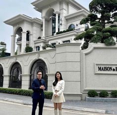 a man and woman standing in front of a large white building with trees on it