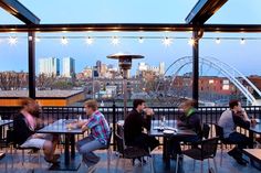 people are sitting at tables in an outdoor dining area with lights strung over the city skyline