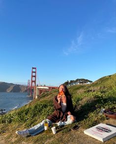 a woman sitting on the ground eating pizza and drinking beer by the golden gate bridge