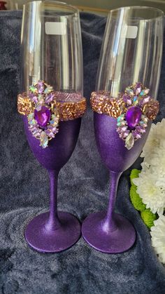 two wine glasses decorated with purple and gold glitters are sitting next to white flowers