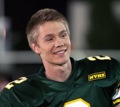 a young man wearing a green and yellow football uniform smiles at the camera while standing in front of an audience