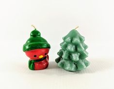 two small christmas trees sitting next to each other on a white surface with one candle in the shape of a snowman