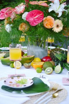 an arrangement of fruit, flowers and drinks on a white table cloth with place settings