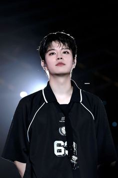 a young man wearing a black shirt and tie with white lettering on the chest, standing in front of a spotlight