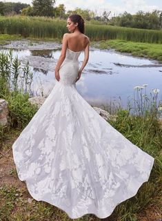 a woman in a white wedding dress standing on the grass next to a pond and looking off into the distance