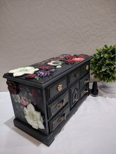 a black dresser with flowers painted on it's drawers and knobs sitting next to a potted plant