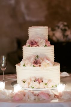 a white wedding cake with pink flowers and candles on the table next to wine glasses