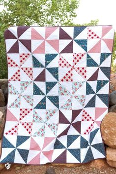 a quilt is displayed on top of some rocks in the background, with trees and rocks behind it