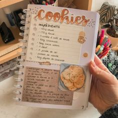 a hand holding up a recipe book with cookies on the page and writing in it