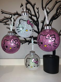 three hello kitty ornaments hanging from a tree with black branches and pink glitter balls on them