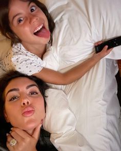 two women laying in bed and one is holding a remote control while the other looks up