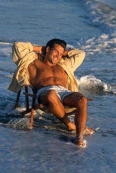 a man sitting in a chair on the beach with his arms around him and smiling