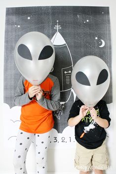 two children standing next to each other with balloons in front of their faces and an alien drawing on the wall behind them