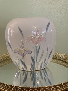 a white vase with flowers painted on it sitting on a glass table next to a gold rimmed platter