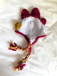 a crocheted chicken hat laying on top of a white bed sheet with yellow and red yarn