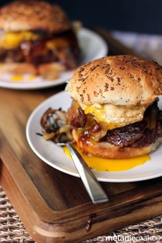 two burgers with bacon and egg on a wooden tray next to a knife and fork