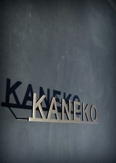 the name kaneeiko is cut out from metal letters on a wall with a shadow