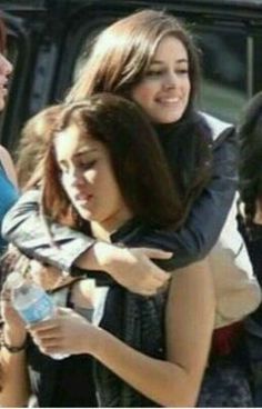 two girls hugging each other while standing in front of a car with another girl looking at her phone