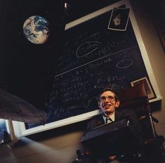 a man sitting in a chair next to a blackboard with writing on it and an image of the earth above him