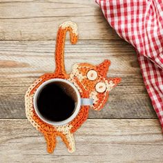 an orange crocheted cat coaster sitting on top of a wooden table next to a cup of coffee