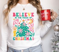 a woman holding a red coffee mug in front of a christmas tree with believe in the magic of christmas written on it