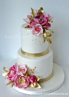 a three tiered white cake with pink flowers on top and gold trimmings