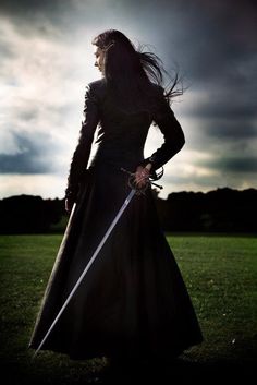 a woman in a long black dress holding two swords and standing on grass with cloudy sky behind her