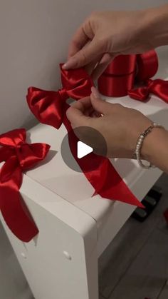 someone is wrapping red ribbon around a white box with bows on it and another person's hand reaching for the bow