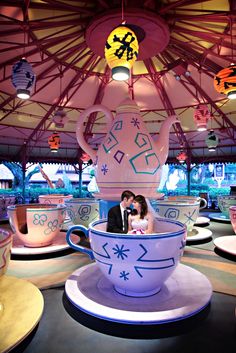 a bride and groom are sitting in a teacup at the fairground, surrounded by brightly lit lights