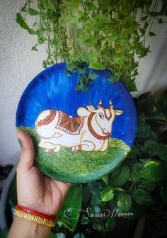 a hand holding up a painted plate with a cow on it's side and green plants in the background