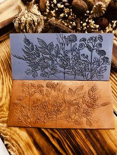 two cards with flowers on them sitting next to some rocks and dried plants in the background