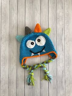 a blue crocheted hat with an orange and green monster on the front, sitting on top of a wooden floor