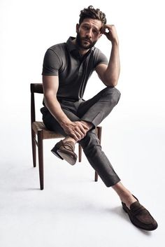 a man sitting on top of a wooden chair next to a white wall and holding his hand near his head