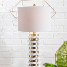 a white and gold lamp on a table in front of a brick wall with a green plant