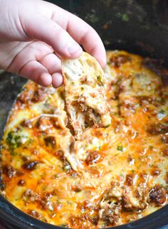 a hand holding a piece of pizza in a crock pot with meat and cheese