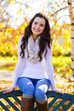 a beautiful young woman sitting on top of a wooden bench in the fall leaves, smiling at the camera