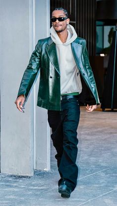 a man in a green leather jacket and sunglasses walking down the street with his hands in his pockets