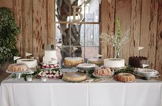 a table topped with lots of cakes and desserts next to a wooden wall in front of a window