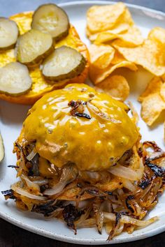 a white plate topped with a cheeseburger and potato chips next to pickles