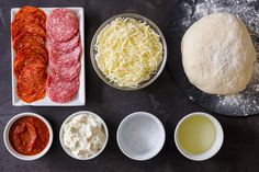 ingredients for pizza laid out in bowls on a black table with flour, cheese and sauces