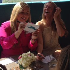 two women sitting at a table laughing and holding papers with a mustache drawn on them