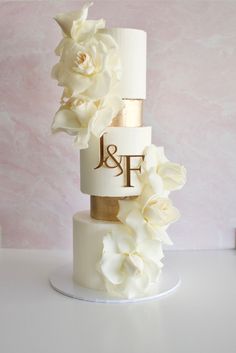 a three tiered white and gold wedding cake with flowers on the top, in front of a pink wall