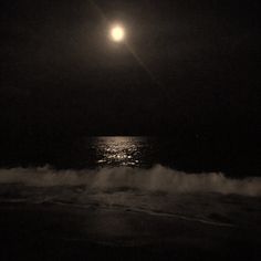 the moon is shining over the ocean on a dark night with waves crashing in front of it