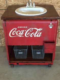 an old coca - cola cooler with two baskets underneath it and a sink on top