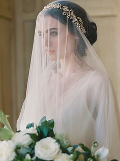 a woman wearing a veil with flowers in the foreground and behind her is a bouquet of white roses