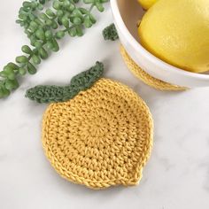 a crocheted lemon sits next to a bowl of lemons and green leaves