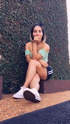 a young woman sitting on the ground next to a hedge with her hand under her mouth