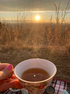 a person holding a cup of tea in front of the sun set over a field