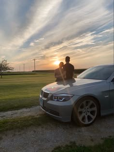 two people sitting on the hood of a silver car in front of a grassy field