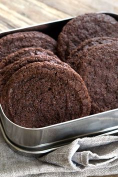 chocolate cookies in a metal container on a wooden table next to a cloth and napkin